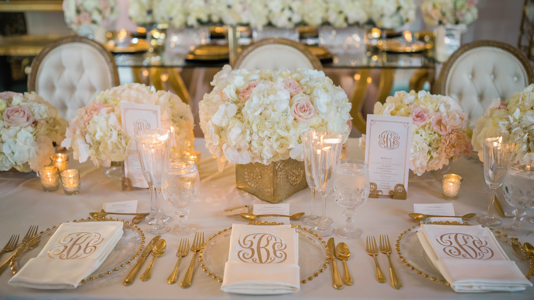 Elegant wedding table setting with gold monogrammed napkins, fine glassware, and floral centerpieces in soft white and blush tones.